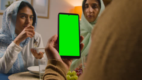 Man-Looking-At-Green-Screen-Mobile-Phone-Sitting-Around-Muslim-Family-Table-At-Home-Eating-Iftar-Meal-Breaking-Daily-Fast-During-Ramadan-1